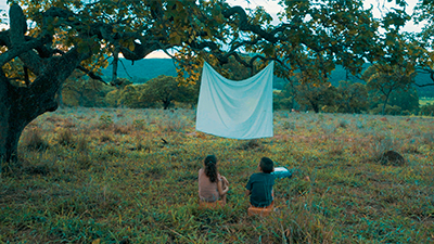 Fotografia de uma cena do filme Quatro bilhões de infinito, de M Marco Antônio Pereira, MG, 2020, Ficção, 15 min. Num campo verde, Adalberto, um garoto de pele branca e cabelos curtos está ao lado da irmã, ela tem pele branca, cabelos castanhos preso por um rabo de cavalo. Eles estão de costas, sentados sobre um bloco de barro, contemplando um lençol branco projetado nas galhas de uma árvore.