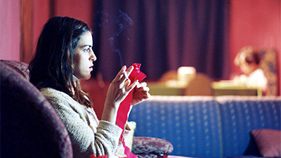Fotografia de uma cena do filme A mão que afaga, de Gabriela Amaral Almeida, SP, 2012, FICÇÃO, 19 min. Sentada numa poltrona roxa, com estampa cinza, uma mulher jovem, branca, de perfil direito, de cabelos lisos pretos até os ombros, usa um pulôver bege, segura nas mãos um papel celofane pink e um cigarro, na mão direita. Ao fundo desfocados, um sofá azul, atrás uma criança à frente de uma mesa, de perfil esquerdo e depois, uma cortina roxa e a parede pink.