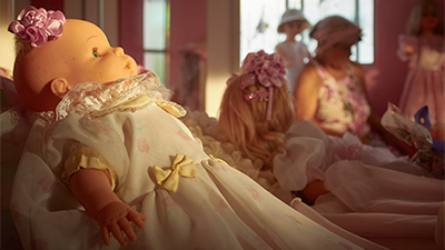 Fotografia de uma cena do filme Açucena, de Isaac Donatto. BA, 2021, DOCUMENTÁRIO, 71 min. Num quarto rosa e branco, com flores e laços rosas e um lilás, aqui na frente, de perfil esquerdo, deitada, uma boneca meu bebê, tem uma flor de cetim rosa, preso no pouquinho de cabelo. Ela usa uma vestido branco, de florzinhas rosas e laço amarelo e gola, com babado com fita rosa. Mais atrás, outras bonecas também de vestidos brancos e rosa e entre elas, uma senhora sentada, com um chapéu e blusas com florais também branco e rosa. O sol suave, atravessa da direita para a esquerda, iluminando a boneca e a senhora, apenas dos ombros para baixo.