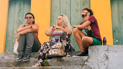 Fotografia de uma cena do filme E o que a gente faz agora? De Marina Pontes, BA, 2019, Ficção, 16 min. Na rua, três jovens sentados na escada à frente de um casarão colonial de parede amarela e porta verde. À esquerda, uma garota de boné preto com aba para trás e óculos escuros está com os braços cruzados sobre as pernas. Ao centro, uma jovem com cabelos loiros, encaracolados, ajeita a alça da blusa e olha para o lado distraidamente. À direita um jovem negro, cabelo black, barbudo, olha para o céu e contrai o rosto diante da intensidade do sol.