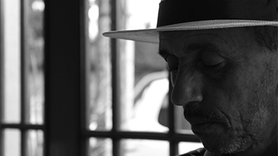 Fotografia de uma cena do filme J. C. D’Almeida, uma foto-síntese, de Rogério Luíz Oliveira, Bahia, 2015, DOCUMENTÁRIO, 26 min. Em preto e branco, sombreado, J. C. D’Almeida, no canto, de perfil esquerdo, visto do queixo para cima, tem a pele clara, e usa chapéu panamá. A face tem muitas rugas, profundas, os olhos fechados, nariz pontiagudo, lábios afinados, bigode e barba rasa, grisalha. Ao fundo, desfocado, um gradeado com visibilidade para rua ensolarada.
