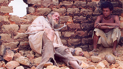 Fotografia do filme Na terra do sol, de Lula Oliveira, BA, 2005, FICÇÃO, 12 min. Dois homens nos escombros da igrejinha de Canudos. Um deles está recostado na parede, sem reboco e com um buraco à esquerda. É um senhor, branco, calvo e com cabelos na altura dos ombros, barba e bigode grisalhos, está com os olhos fechados. Usa uma túnica bege e uma calça cinza, ambos esfarrapados e uma sandália. Ele segura uma cruz, feita de galhos. À direita, sentado num monte de terra, um jovem negro, de cabelos black, pretos, usa uma bermuda jeans rasgada. Está com os braços apoiados sobre os joelhos e olha misericordioso para o senhor.