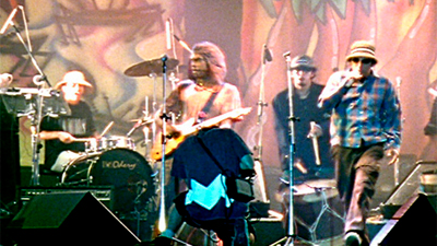 Fotografia de uma cena do filme O mundo e uma cabeça, de Bidu Queiroz, Cláudio Barroso PE, 2005, DOCUMENTÁRIO, 17 min. Quatro integrantes da banda Chico Science & Nação Zumbi estão num palco. Da esquerda para a direita, o baterista, sentado, um homem pardo, com chapéu bege e camiseta preta; ao lado dele, o guitarrista, em pé, usa um gorro e uma camiseta marrom clara; um pouco atras, o terceiro toca um tambor de Alfaia, usa um pequeno chapéu arredondado, preto e casaco e calça, pretos. À direita, Chico Science, é um homem negro não retinto, usa um chapéu bege, com faixas coloridas, uma camisa de manga comprida, xadrez, azul e vermelha, calça marrom e tênis. Segura um microfone. Ao fundo, grafite em tons vermelho, marrom terra, amarelo, verde e preto, com desenhos de palmeiras, um caranguejo e traços curvilíneos.