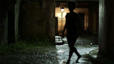 Sombreada na contraluz, a direita, a silhueta de uma mulher caminha numa rua de paralelepípedos, ela tem cabelo black. Ao fundo, numa casa de portões de ferro, uma luminária colonial, clareia este lado da rua, por ela passa. À esquerda, paredes grossas, desgastadas e vegetação no chão.