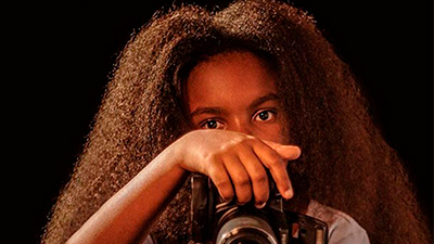 Uma jovem negra retinta, dos ombros para cima, de cabelos crespos, longos, castanhos escuros. Ela tem a testa larga, sobrancelhas grossas, olha para nós por sobre a mão à esquerda, que empunha uma câmera fotográfica profissional. Usa uma camiseta azul clara.