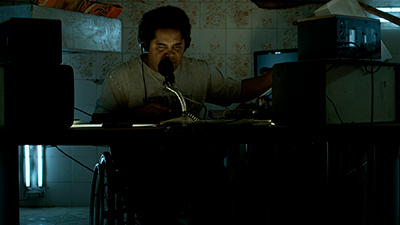 Num cômodo de azulejos florais amarelados, um homem negro, de cabelos curtos, pretos, crespos, olhar contraído, nariz largo, camiseta bege e fone de ouvidos grandes, está à frente de uma mesa, com um microfone. Ele tem o braço à direita com a mão mexendo nos equipamentos de som deste lado. Do outro lado, outras duas caixas, estão escurecidas pela pouca luz, que ilumina mais o rosto dele. Embaixo da mesa, ao fundo, à esquerda, duas luminárias verticais, clareiam a cadeira de rodas que ele senta.