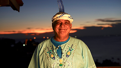 À frente da cidade, durante um por do sol, de céu azul, uma faixa alaranjada e a abaixo, no escuro da noite, luzes de carros e prédios, Gerônimo. Ele usa um turbante branco e azul, enrolado na testa, com uma pena azul presa na parte de trás. Ele é negro não retinto, gordo, o sorriso deixa os olhos encolhidos, usa uma bata verde clara, com bordados no peito, sobre uma camiseta azul.