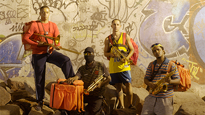 Quatro rapazes, com bolsas de entregador laranja seguram instrumentos musicais metálicos de sopro