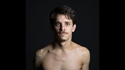 À frente de um fundo preto, um homem branco, do peito para cima. Ele é branco, tem os cabelos castanhos escuros, as sobrancelhas e o bigode castanhos claro, os lábios rosa, cerrados e os olhos focados na gente, caramelo. No ombro à direita, tem uma tatuagem tribal. À esquerda, parte do rosto está sombreado.