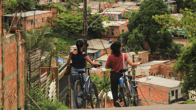 No alto de uma ladeira, duas mulheres, de costas seguram uma bicicleta azul cada uma, do lado direito. As duas são brancas e magras. A da esquerda é branca, tem o cabelo liso, preso num rabo de cavalo, usa uma blusa azul marinho, calça jeans e uma bolsa marrom. A outra tem o cabelo ondulado, preto, pouco abaixo dos ombros, preso na parte cima; usa uma blusa vermelha, calça e tênis preto e uma ecobag branca. Ao fundo e abaixo, uma comunidade de casas de alvenaria, sem reboco, entremeadas por árvores, um poste e fiação elétrica.