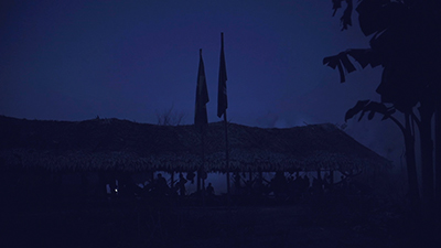 Fotografia de uma cena do filme. Na penumbra noturna, em uma alongada Oca, uma família indigena, em redes e em pé, está reunida. Uma intensa fumaça encobre, acima e o entorno. À direita, algumas palmeiras e aqui na frente duas bandeiras, uma do Brasil.