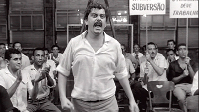 Fotografia em preto e branco, de uma cena do filme. Num galpão, com paredão com janelas pequenas ao fundo, diversos homens sentados, a maioria de camisa social, de manga curta branca e calça, aplaudem e observam um homem que está em pé, bem à frente, no meio. Ele é pardo, tem cabelos curtos encaracolados, tem bigode, usa camisa social, branca de manga comprida arregaçada até os cotovelos e calça social cinza. Ele tem o olhar serio e está com a boca aberta.