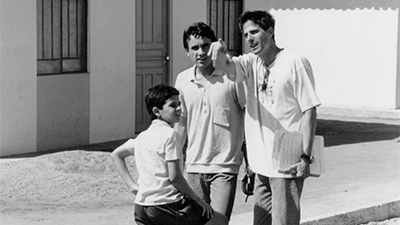 Fotografia em preto e branco de uma cena do filme. Numa manhã de sol, dois jovens e um menino, em frente a uma casa. Os três são brancos e tem cabelos curtos escuros. O mais alto, à direita, tem cabelos lisos, usa uma bata clara, um óculos escuros pendurados na bata, está de calça, segura papéis ao lado corpo e aponta para frente. À esquerda deste, o outro é um pouco menor, tem os cabelos ondulados, olha para nós, está com uma camisa pólo clara, calça e segura um cigarro na mão esquerda. Logo ao lado dele, o menino, de costas para nós, olha para a direita, com as sobrancelhas contraídas. Está com camisa de botão, de manga curta, branca e um shorts preto. Está com a mão esquerda na cintura e a outra apoiada na perna.