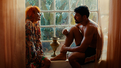 Fotografia de uma cena do filme. Sentados no parapeito de uma grande janela de vidro, na contraluz, uma senhora e um jovem, voltados um para o outro. Helena (Helena Ignez), à esquerda é branca, tem os cabelos lisos, laranja, na altura do ombro, com franja, usa um vestido de mangas compridas, colorido com ênfase laranja. Ela tem as mãos apoiadas sobre as coxas e olha para ele, serena sorridente. Alexandre (Cauã Raymond), é branco, tem os cabelos curtos, escuros, usa bigode e barba curtos, bronzeado, está de shorts preto, com faixa vermelha e branca na lateral. Ele tem a perna direita sobre o parapeito, e está abraçado ao joelho. Olha para baixo. Entre eles um vaso de cobre, ao fundo a vegetação e a luz, adentra iluminando os dois e as cortinas salmão transparente.