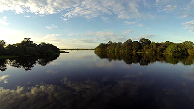 Fotografia de uma cena do filme. Um rio largo aqui na frente e afunilado ao fundo pela vegetação nas laterais, tem refletido no espelho d’água, o céu azul claro, com nuvens esparsas e a suave luz do sol mais intensa à esquerda. O horizonte é uma fina faixa de terra distante.
