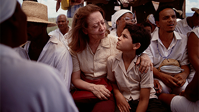 Fotografia de uma cena do filme. Sentados, Dora e Josué (Fernanda Montenegro e Vinicius de Oliveira) se olham intensamente, com as sobrancelhas contraídas. Estão entre várias pessoas negras, vestidas de branco, duas mulheres com lenços, um homem com chapéu de palha, outro com uma conta azul e branca. Os dois são brancos. Dora tem os cabelos lisos, ruivos, na altura dos ombros, está com uma camisa de botão, bege, de manga curta, uma calça vinho e segura a bolsa sobre o colo. Josué, Tem os cabelos curtos, lisos, pretos, usa uma camisa de botão, acizentada, de manga curta e um shorts preto. Ele está recostado nela, que o abraça no ombro.