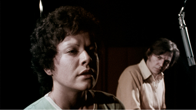 Fotografia de uma cena do filme. Elis Regina e Tom Jobim, jovens. Aqui na frente, à esquerda, Elis, a partir do ombro. Ela é branca, está com os cabelos curtos, ondulados, escuros, tem a testa contraída e os olhos pequenos, encolhidos, voltados para a direita, distantes. Usa uma camiseta verde clara. Ao fundo, quase de perfil, da cintura para cima, Tom Jobim com a cabeça inclinada, olha para ela. Ele é branco, tem os cabelos lisos, castanhos claro, quase no ombro. Usa uma camisa social bege. Acima da cabeça dele, um microfone.