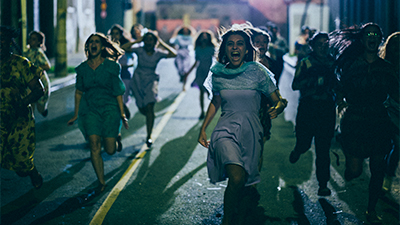 Fotografia de uma cena do filme. Numa rua, à noite, diversas mulheres correm em nossa direção gritando. As do meio usam vestidos azuis e sandálias. Uma delas está com as mãos na cabeça. Muitas delas têm cabelos lisos, abaixo do ombro. Uma luz noturna é irradiada do fundo para frente.